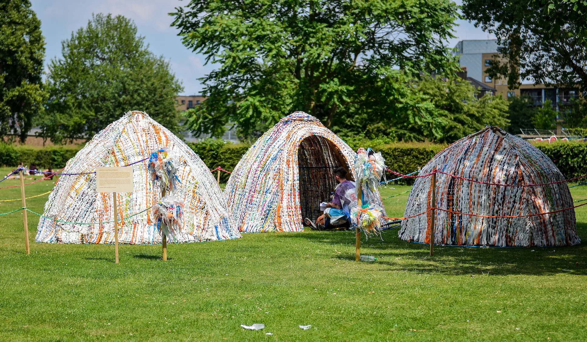 Three domed huts woven from coloured plastic strips on a park lawn, roped off, with a visitor sitting in the doorway of one