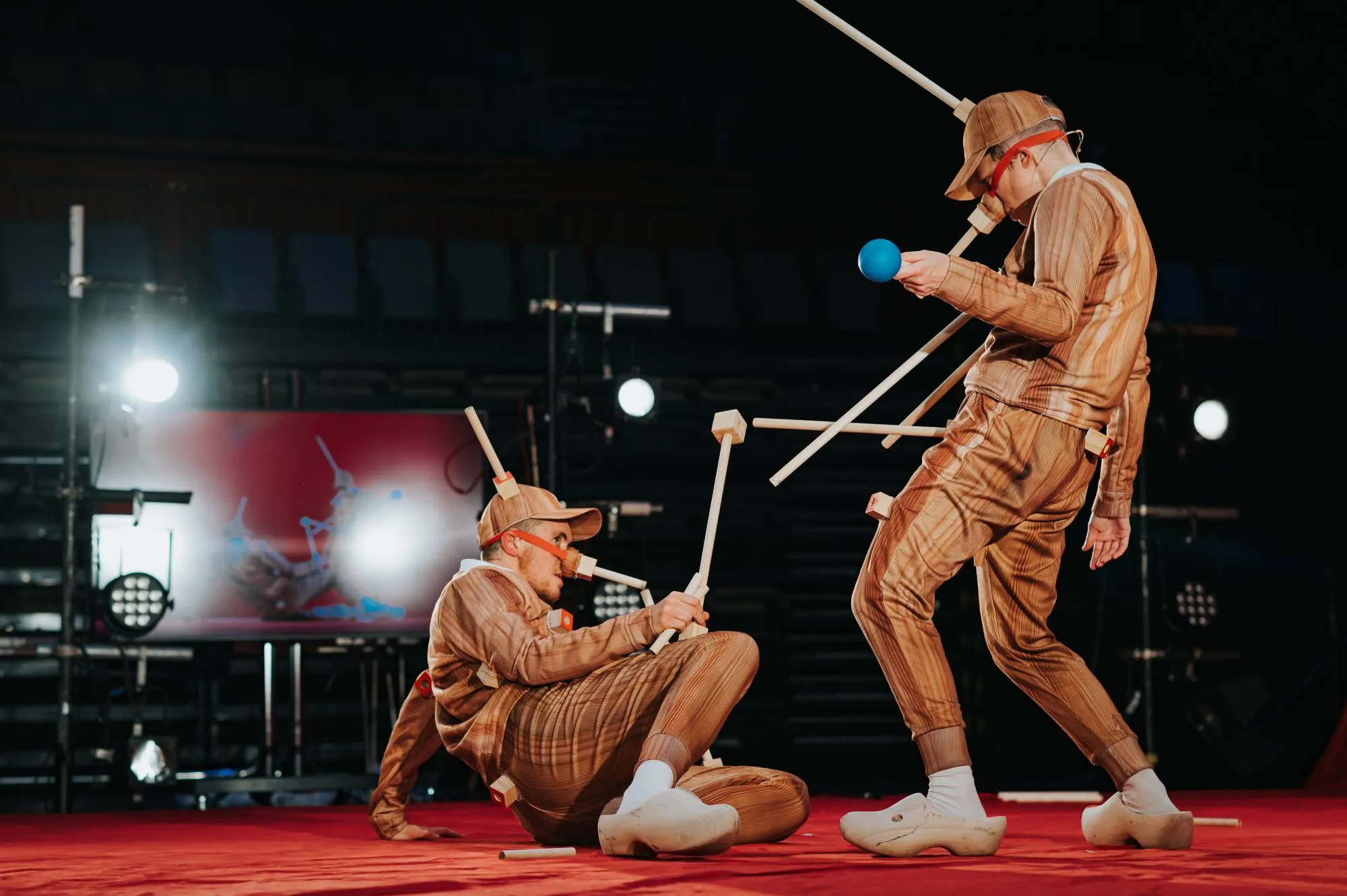 Ivor MacAskill and Rosana Cade on a dark stage in fake-wooden tracksuits, with wooden appendages protruding from their heads and noses