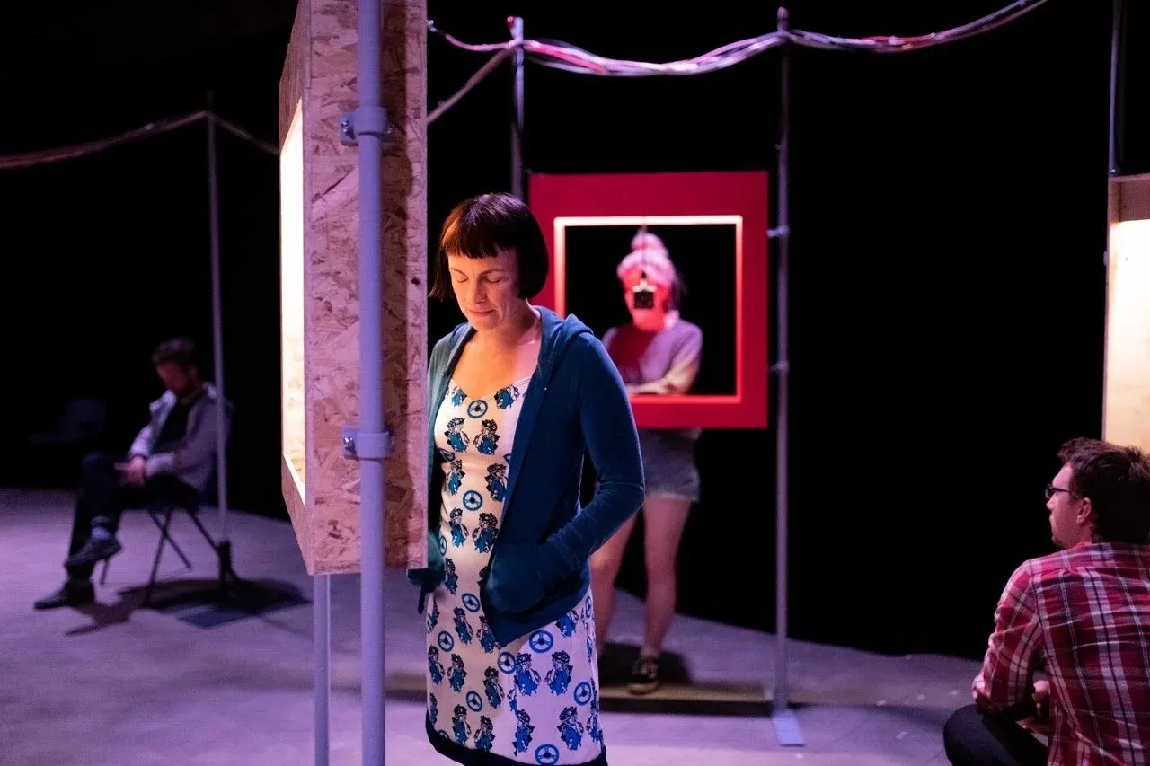 A person with eyes closed stands among plywood booths in a darkened gallery, listeners seated nearby